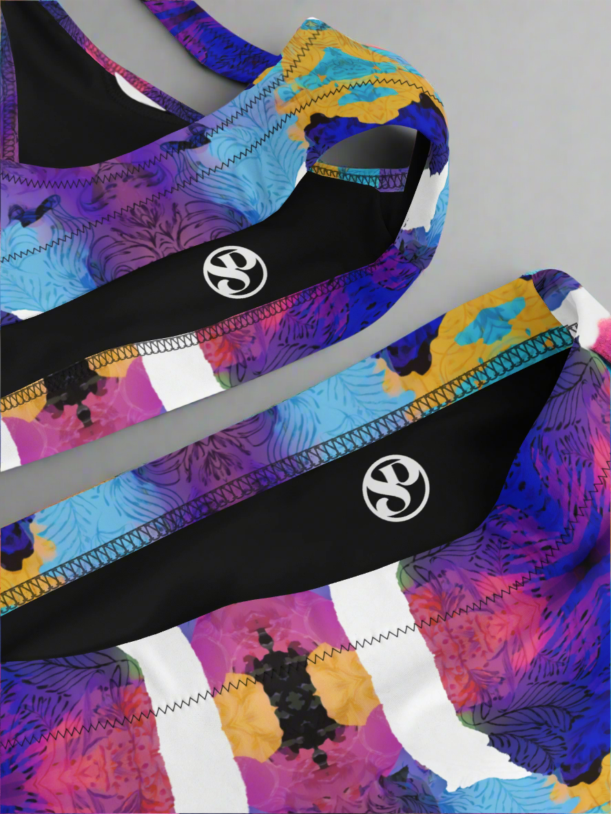 Close-up flat lay of Pixie & Sherbet “Ocean Mirage Surf” bikini — vibrant abstract print in purple, blue, yellow, black, and white, featuring sports-bra style top and matching high-waisted bottoms with detailed stitching, displayed on a plain grey background.