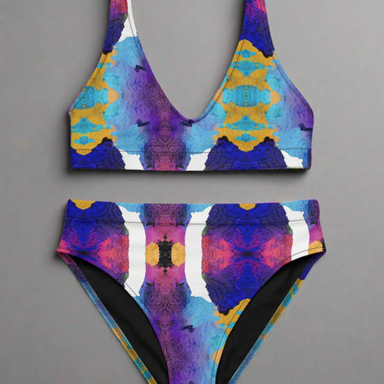 Front view flat lay of Pixie & Sherbet “Ocean Mirage Surf” bikini — vibrant kaleidoscopic print in blue, purple, pink, yellow, and white, featuring deep V-neck top with wide straps and high-waisted bottoms, displayed on a plain grey background.