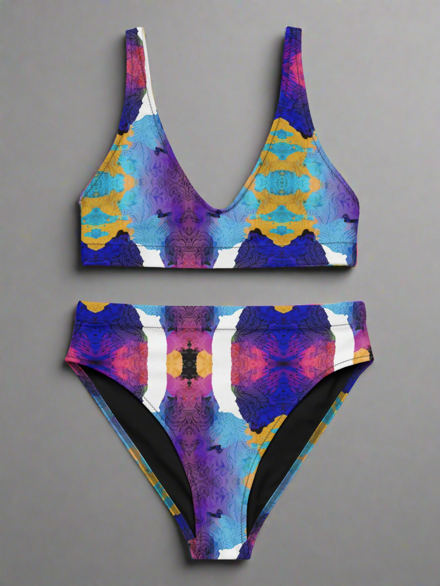 Front view flat lay of Pixie & Sherbet “Ocean Mirage Surf” bikini — vibrant kaleidoscopic print in blue, purple, pink, yellow, and white, featuring deep V-neck top with wide straps and high-waisted bottoms, displayed on a plain grey background.