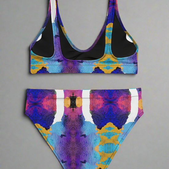 Back view flat lay of Pixie & Sherbet “Ocean Mirage Surf” bikini — vibrant kaleidoscopic print in purple, blue, yellow, pink, and white, featuring scoop-neck sports-bra style top with wide straps and high-waisted bottoms, displayed on a plain grey background.