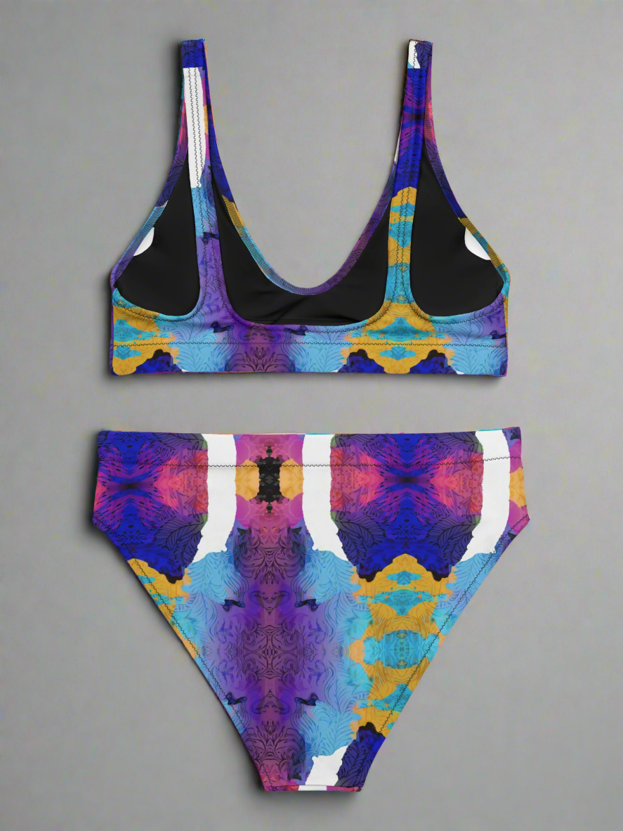 Back view flat lay of Pixie & Sherbet “Ocean Mirage Surf” bikini — vibrant kaleidoscopic print in purple, blue, yellow, pink, and white, featuring scoop-neck sports-bra style top with wide straps and high-waisted bottoms, displayed on a plain grey background.