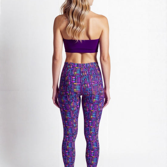 Back view of a woman wearing Pixel Pulse Crossover Leggings with a vibrant geometric pattern in purple, blue, and pink, paired with a purple sports bra and white sneakers, standing against a plain background.