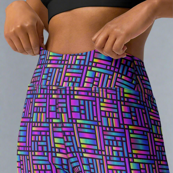 Woman adjusting waistband of Pixel Pulse Yoga leggings with vibrant multicolored geometric pattern in purple, blue, green, and pink, paired with a black crop top, against a neutral background