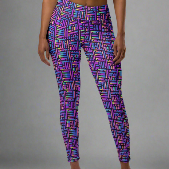 Front view of woman wearing Pixel Pulse Yoga leggings with vibrant multicolored geometric pattern in purple, pink, blue, green, and yellow, paired with a black sports bra, barefoot against a plain gray background.
