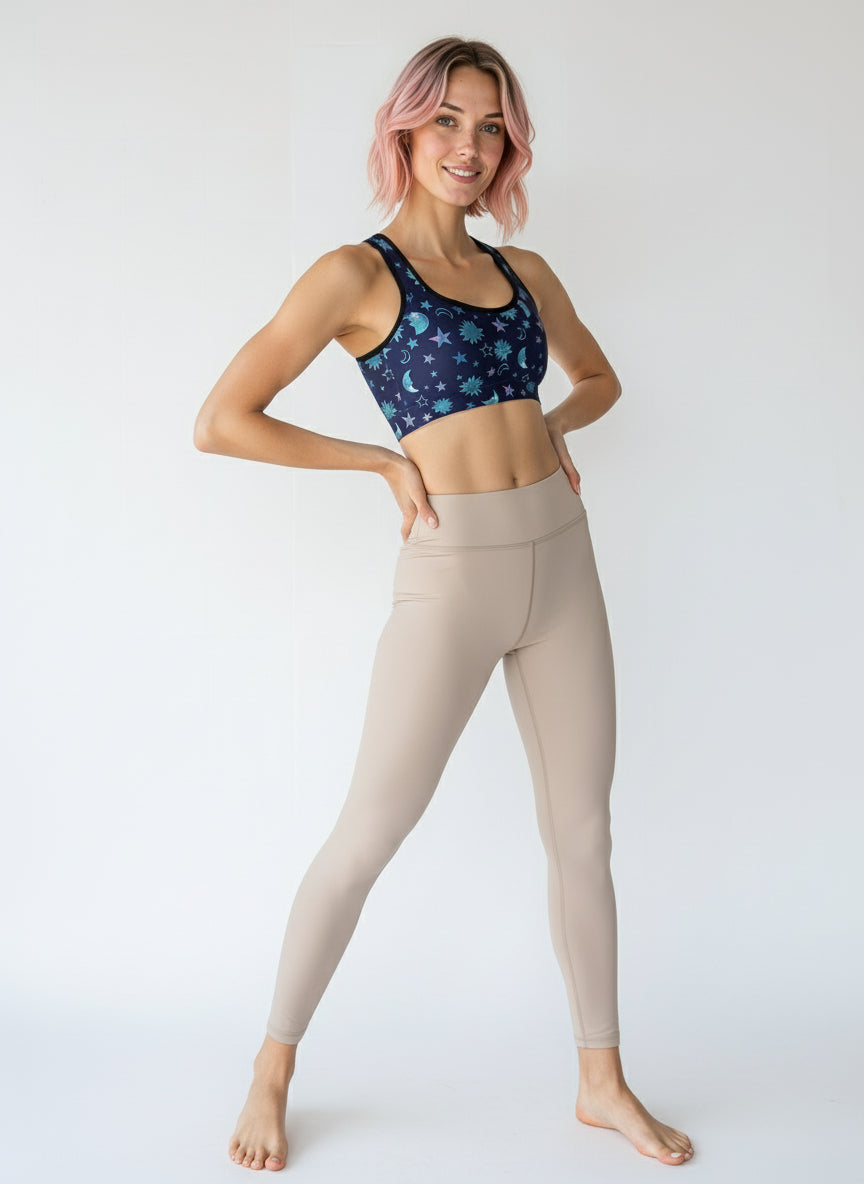 Woman wearing Cosmic Calm Essential Active Bra with a navy base and pastel blue and purple celestial print, smiling and standing against a plain background