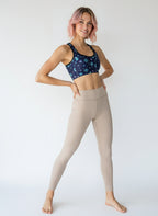 Woman wearing Cosmic Calm Essential Active Bra with a navy base and pastel blue and purple celestial print, smiling and standing against a plain background