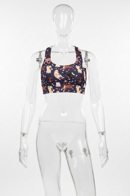 A women's sports bra with a navy blue background and an all-over print of various dog breeds in beige, brown, and black, interspersed with small white bone and paw print motifs. It features a white racerback-style back. 