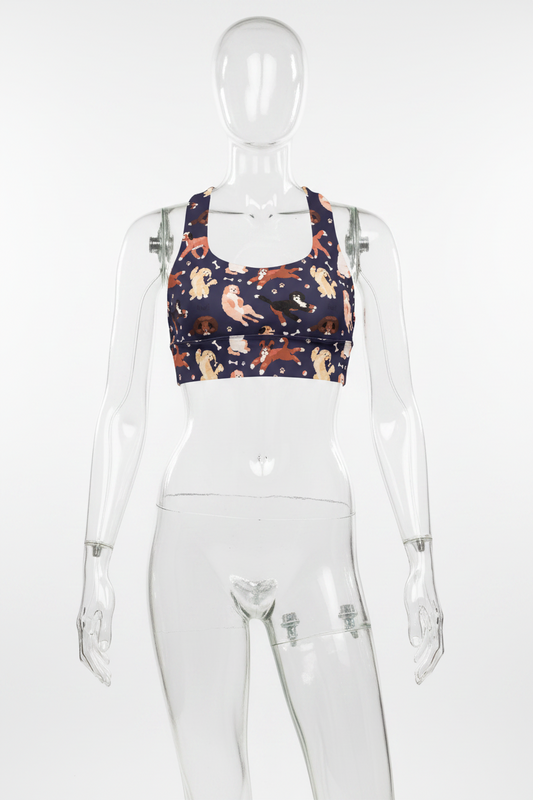A women's sports bra with a navy blue background and an all-over print of various dog breeds in beige, brown, and black, interspersed with small white bone and paw print motifs. It features a white racerback-style back. 