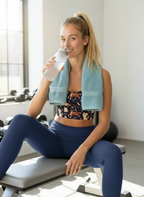 Woman sitting on a gym bench with a towel over her shoulder and a water bottle, smiling.