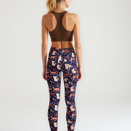 A woman is seen from behind wearing Puppy Parade crossover leggings with a high-waisted fit and an all-over print of cartoon puppies and bones in warm tones on a navy background. She pairs the leggings with a brown racerback sports bra and white sneakers, standing against a plain, light backdrop.