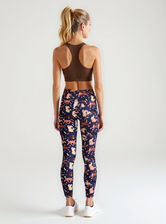 A woman is seen from behind wearing Puppy Parade crossover leggings with a high-waisted fit and an all-over print of cartoon puppies and bones in warm tones on a navy background. She pairs the leggings with a brown racerback sports bra and white sneakers, standing against a plain, light backdrop.