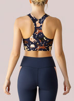 A close-up of a woman’s back showing the Puppy Parade longline bra, featuring a racerback design and a playful print of cartoon puppies and bones in shades of cream, brown, and black on a navy background. She is also wearing high-waisted black leggings, with a plain, light backdrop.