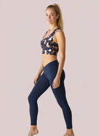 Side view showing the Puppy Parade longline bra, featuring a racerback design and a playful print of cartoon puppies and bones in shades of cream, brown, and black on a navy background. She is also wearing high-waisted black leggings, with a plain, light backdrop.