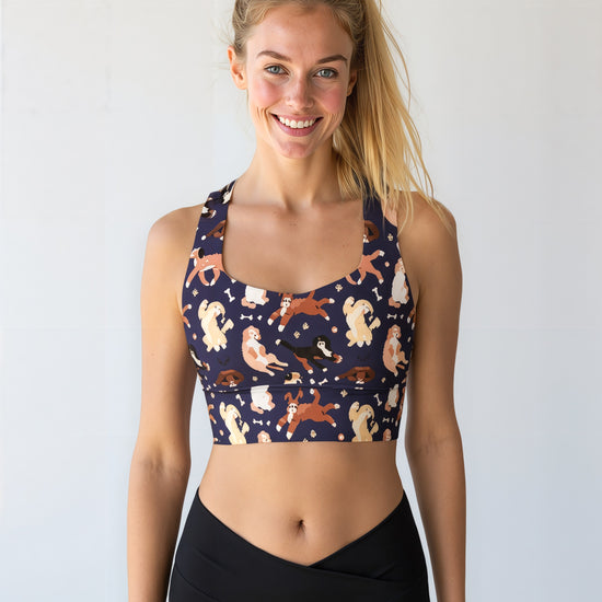 A woman smiles while wearing the Puppy Parade longline bra, featuring a playful print of various cartoon puppies in shades of cream, brown, and tan on a dark navy background. She pairs the bra with high-waisted black leggings and stands against a plain, light background.