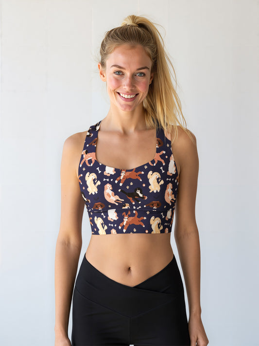 A woman smiles while wearing the Puppy Parade longline bra, featuring a playful print of various cartoon puppies in shades of cream, brown, and tan on a dark navy background. She pairs the bra with high-waisted black leggings and stands against a plain, light background.