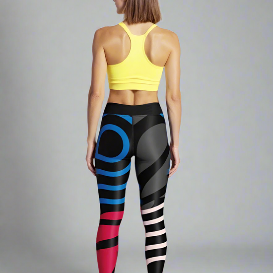 A woman is shown from behind, wearing a yellow sports bra and Red, Blue & Onyx Abstract Yoga Leggings. The leggings feature bold geometric patterns in black, blue, red, and pale pink, creating a dynamic and modern look. She is standing barefoot against a plain, softly lit background, highlighting the unique design of the leggings.