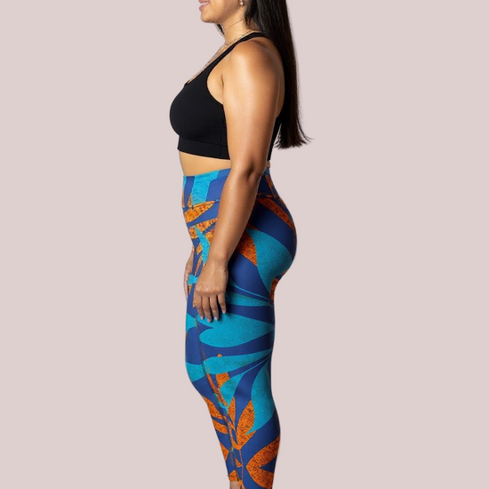 Side profile of a model wearing Pixie & Sherbet’s Rustic Rhythm Yoga leggings with bold blue and orange leaf-inspired patterns, paired with a black sports bra, standing barefoot against a white background in a relaxed, upright pose.