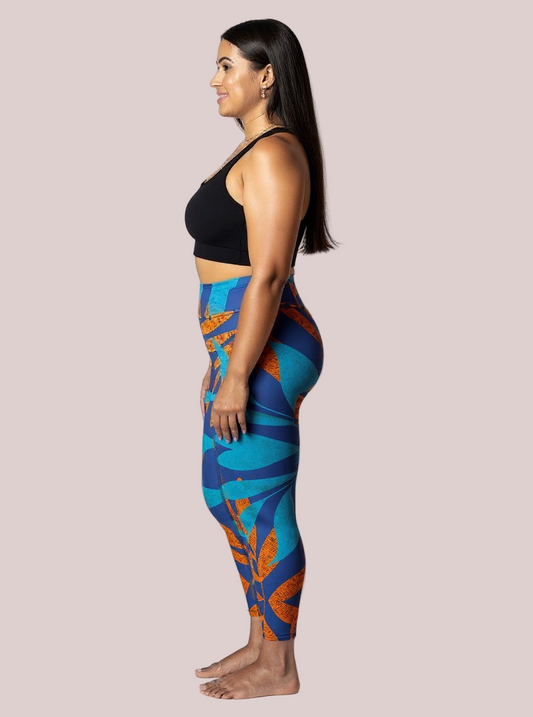 Side profile of a model wearing Pixie & Sherbet’s Rustic Rhythm Yoga leggings with bold blue and orange leaf-inspired patterns, paired with a black sports bra, standing barefoot against a white background in a relaxed, upright pose.