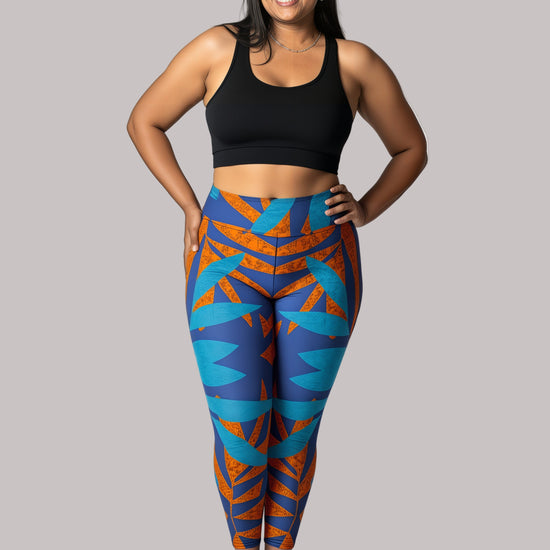Model smiling in Pixie & Sherbet’s Rustic Rhythm Yoga leggings with bold blue and burnt orange geometric patterns, paired with a black sports bra, standing barefoot against a white background in a relaxed, confident pose