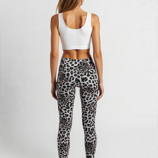 A woman is shown from behind wearing a white sports bra and Savannah Charge Crossover Leggings. The leggings have a grey base with a black and white leopard print pattern and a high-waisted, crossover waistband. She stands barefoot against a plain, softly lit background, showcasing the stylish and comfortable design.