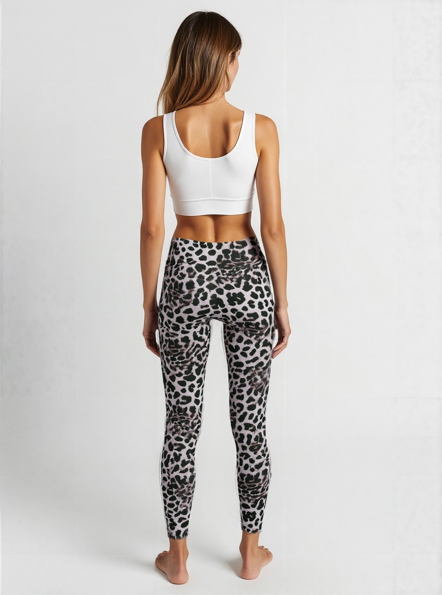 A woman is shown from behind wearing a white sports bra and Savannah Charge Crossover Leggings. The leggings have a grey base with a black and white leopard print pattern and a high-waisted, crossover waistband. She stands barefoot against a plain, softly lit background, showcasing the stylish and comfortable design.
