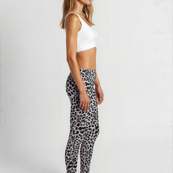 A woman is standing in profile against a plain background, wearing a white sports bra and Savannah Charge Crossover leggings. The leggings feature a stylish black and grey animal print pattern with a flattering high-rise crossover waistband. She is barefoot, and the look showcases a modern, eye-catching activewear style.