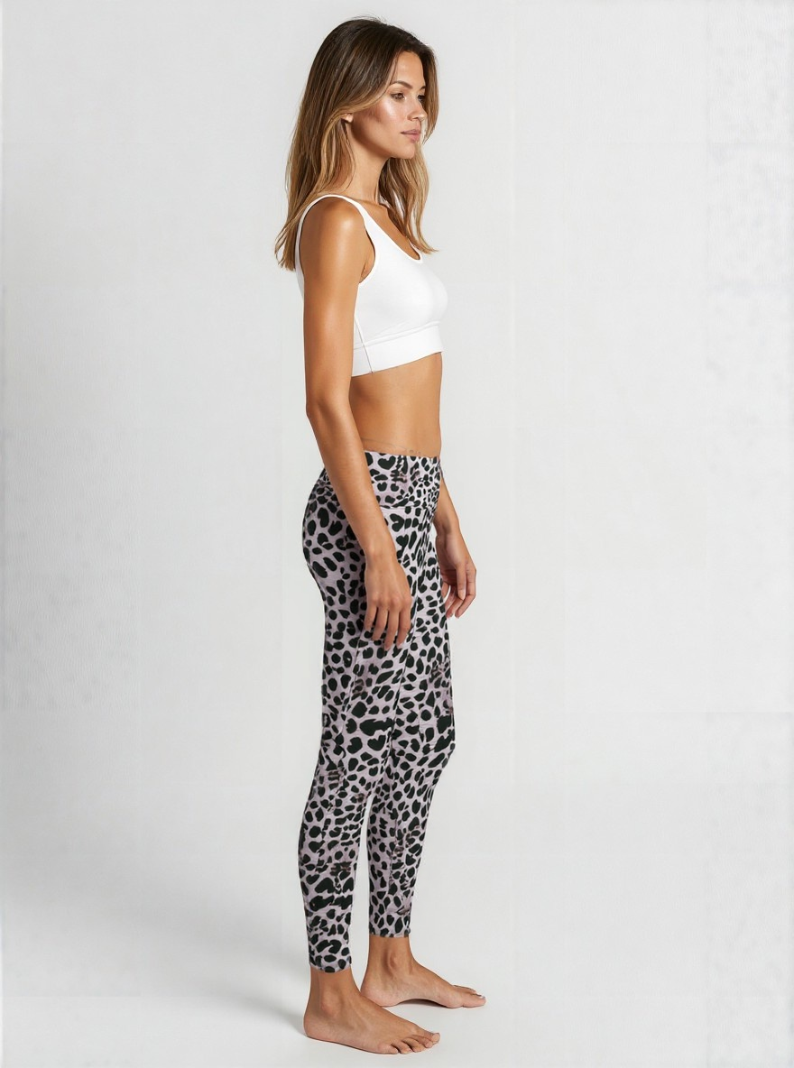 A woman is standing in profile against a plain background, wearing a white sports bra and Savannah Charge Crossover leggings. The leggings feature a stylish black and grey animal print pattern with a flattering high-rise crossover waistband. She is barefoot, and the look showcases a modern, eye-catching activewear style.