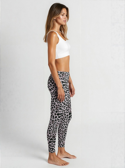 A woman is standing in profile against a plain background, wearing a white sports bra and Savannah Charge Crossover leggings. The leggings feature a stylish black and grey animal print pattern with a flattering high-rise crossover waistband. She is barefoot, and the look showcases a modern, eye-catching activewear style.