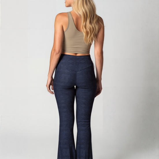 Back view of a woman with long, wavy blonde hair wearing navy blue Shadowleaf flare leggings with a subtle leaf pattern, a fitted beige crop top, and white sneakers, standing against a light background. The image highlights the flared leg and back fit of the leggings.