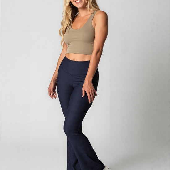 Woman with long, wavy blonde hair smiling and wearing navy blue Shadowleaf flare leggings, a fitted beige crop top, and white sneakers, standing against a light background. The leggings have a subtle leaf pattern and a flared silhouette.