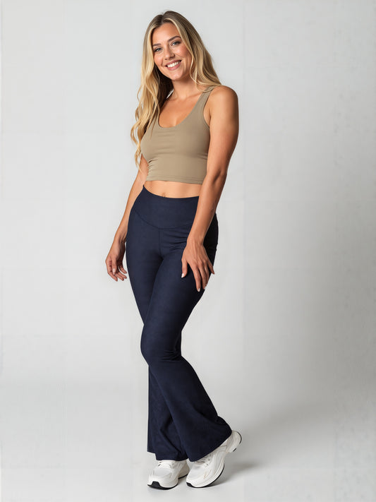 Woman with long, wavy blonde hair smiling and wearing navy blue Shadowleaf flare leggings, a fitted beige crop top, and white sneakers, standing against a light background. The leggings have a subtle leaf pattern and a flared silhouette.