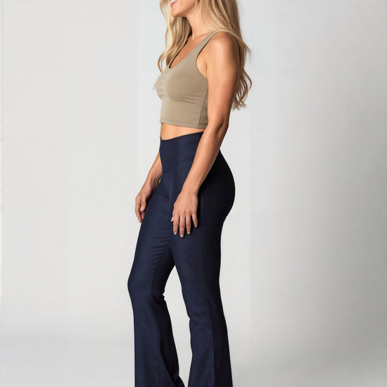 Side view of a woman with long, wavy blonde hair smiling and wearing navy blue Shadowleaf flare leggings, a fitted beige crop top, and white sneakers, standing against a light background. The leggings have a subtle leaf pattern and a flared leg.
