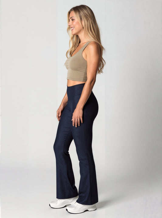 Side view of a woman with long, wavy blonde hair smiling and wearing navy blue Shadowleaf flare leggings, a fitted beige crop top, and white sneakers, standing against a light background. The leggings have a subtle leaf pattern and a flared leg.