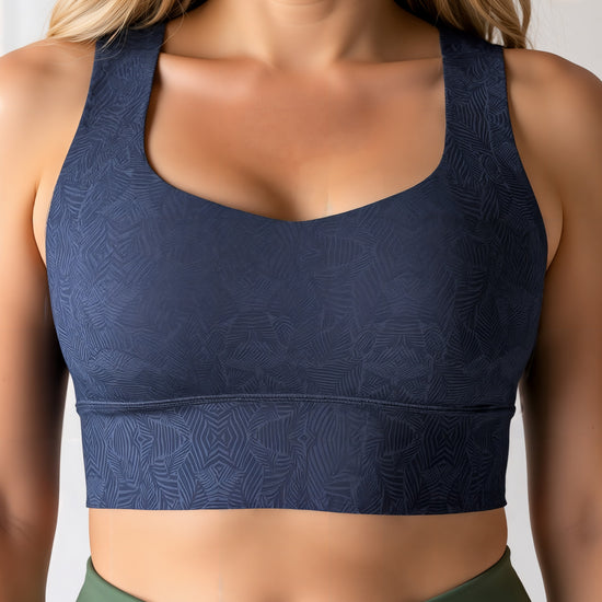 Close-up of a woman wearing a dark blue Shadowleaf longline sports bra with a subtle leaf pattern, paired with high-waisted olive green leggings. The image focuses on her torso, showing the fit and detailing of the sports bra and leggings.