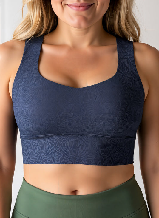 Close-up of a woman wearing a dark blue Shadowleaf longline sports bra with a subtle leaf pattern, paired with high-waisted olive green leggings. The image focuses on her torso, showing the fit and detailing of the sports bra and leggings.