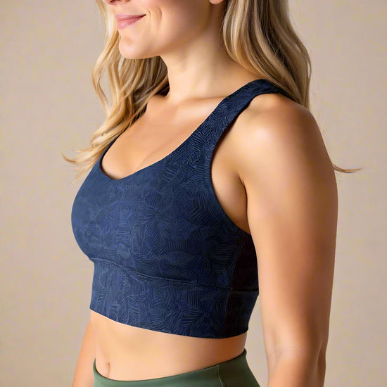 Side view of a woman with long, wavy blonde hair smiling and wearing a dark blue Shadowleaf longline sports bra with a subtle leaf pattern, paired with high-waisted olive green leggings. The image highlights the fit and texture of the sports bra.
