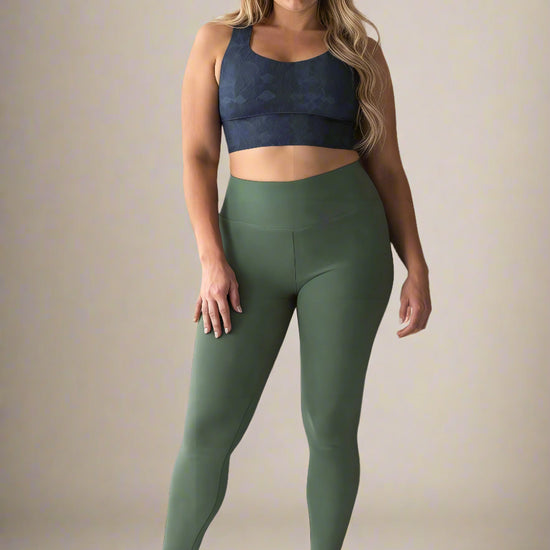 Curvy woman with long, wavy blonde hair wearing a dark blue Shadowleaf longline sports bra and high-waisted olive green leggings, standing barefoot and smiling confidently against a light background.