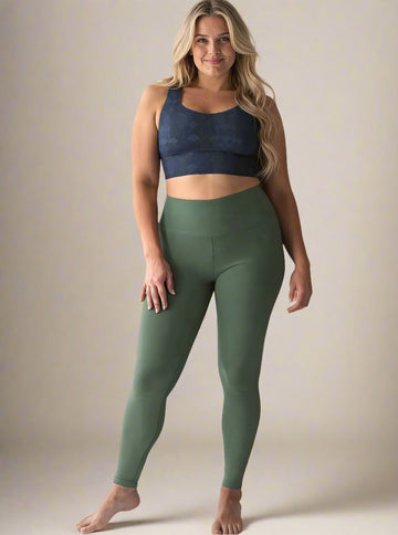 Curvy woman with long, wavy blonde hair wearing a dark blue Shadowleaf longline sports bra and high-waisted olive green leggings, standing barefoot and smiling confidently against a light background.