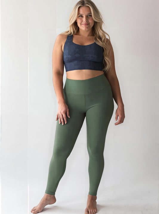 Curvy woman with long, wavy blonde hair wearing a dark blue Shadowleaf longline sports bra and high-waisted olive green leggings, standing barefoot and smiling confidently against a light background.