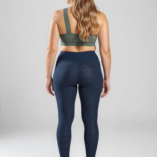 Back view of a woman wearing navy blue high-waisted Shadowleaf Pocket Leggings and a sage green sports bra, standing on a plain light grey background.