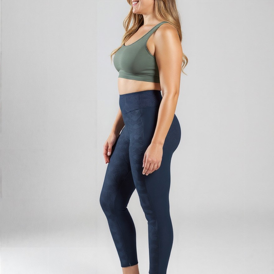 Smiling woman wearing navy blue high-waisted Shadowleaf Pocket Leggings and a sage green sports bra, standing in profile against a light grey background.