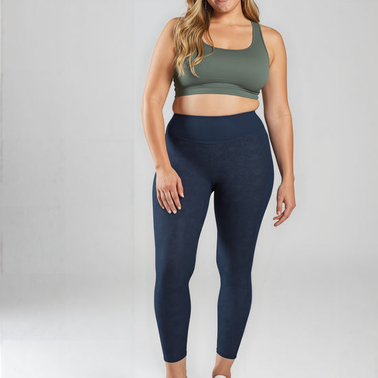 A woman with long, wavy hair is smiling and standing against a light background, wearing an olive green sports bra and high-waisted, dark navy Shadowleaf pocket leggings with subtle botanical patterns and white sneakers.