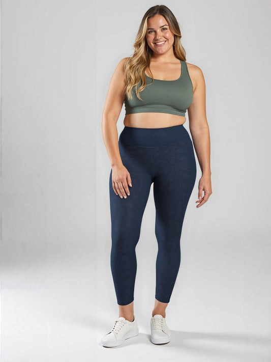 A woman with long, wavy hair is smiling and standing against a light background, wearing an olive green sports bra and high-waisted, dark navy Shadowleaf pocket leggings with subtle botanical patterns and white sneakers.
