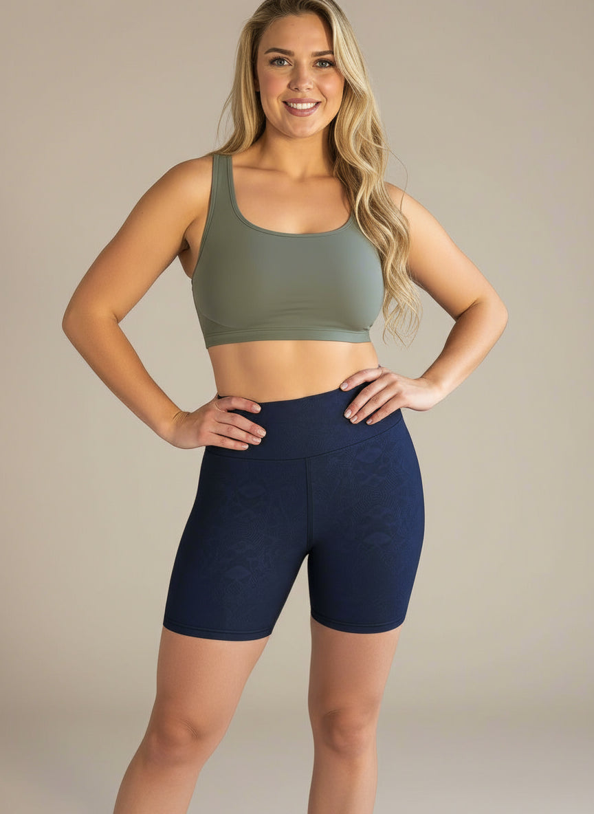  A woman with long blonde hair is smiling and standing against a light background, wearing an olive green sports bra and high-waisted, dark navy Shadowleaf Yoga shorts with subtle textured detailing.