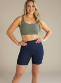  A woman with long blonde hair is smiling and standing against a light background, wearing an olive green sports bra and high-waisted, dark navy Shadowleaf Yoga shorts with subtle textured detailing.