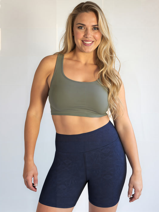  A woman with long blonde hair is smiling and standing against a light background, wearing an olive green sports bra and high-waisted, dark navy Shadowleaf Yoga shorts with subtle textured detailing.