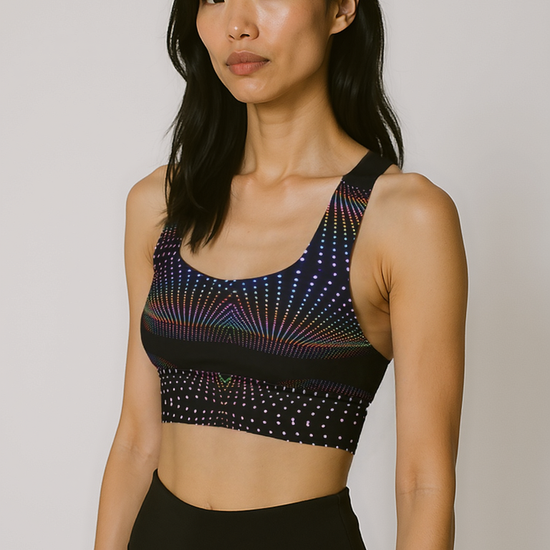 Woman wearing Starlit Tempo longline sports bra with a black base and vibrant, multicolored starburst dot pattern, standing against a plain background.