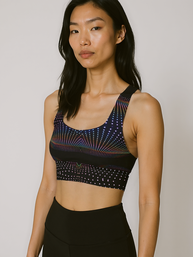 Woman wearing Starlit Tempo longline sports bra with a black base and vibrant, multicolored starburst dot pattern, standing against a plain background.