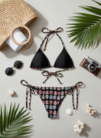 Black triangle bikini with colorful skull pattern bottom, sunglasses, camera, and palm leaves on a light gray background