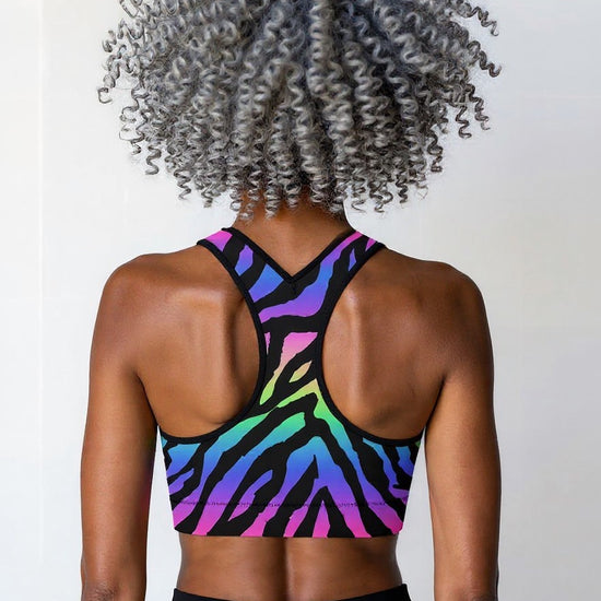 Woman showing the back of the Technicolour Essential Active Bra with bold neon green, blue, and pink zebra stripes, standing against a plain background